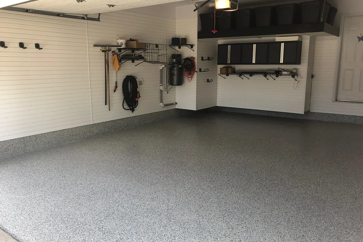 A finished garage with white wall panels, black wall cabinets, an overhead storage rack and a flake-coated floor