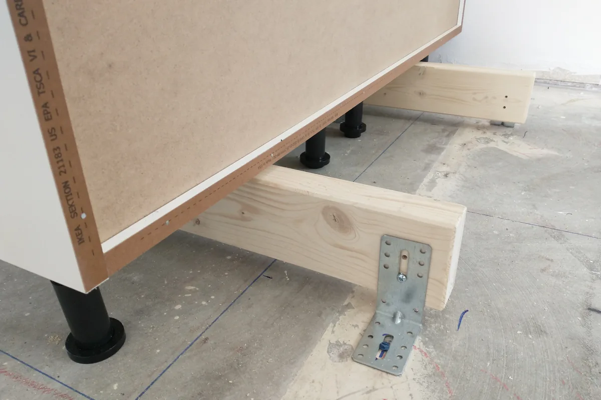 A kitchen island carcass on adjustable legs with a timber cleat and galvanised angle bracket fixed to the floor, Newport Beach CA