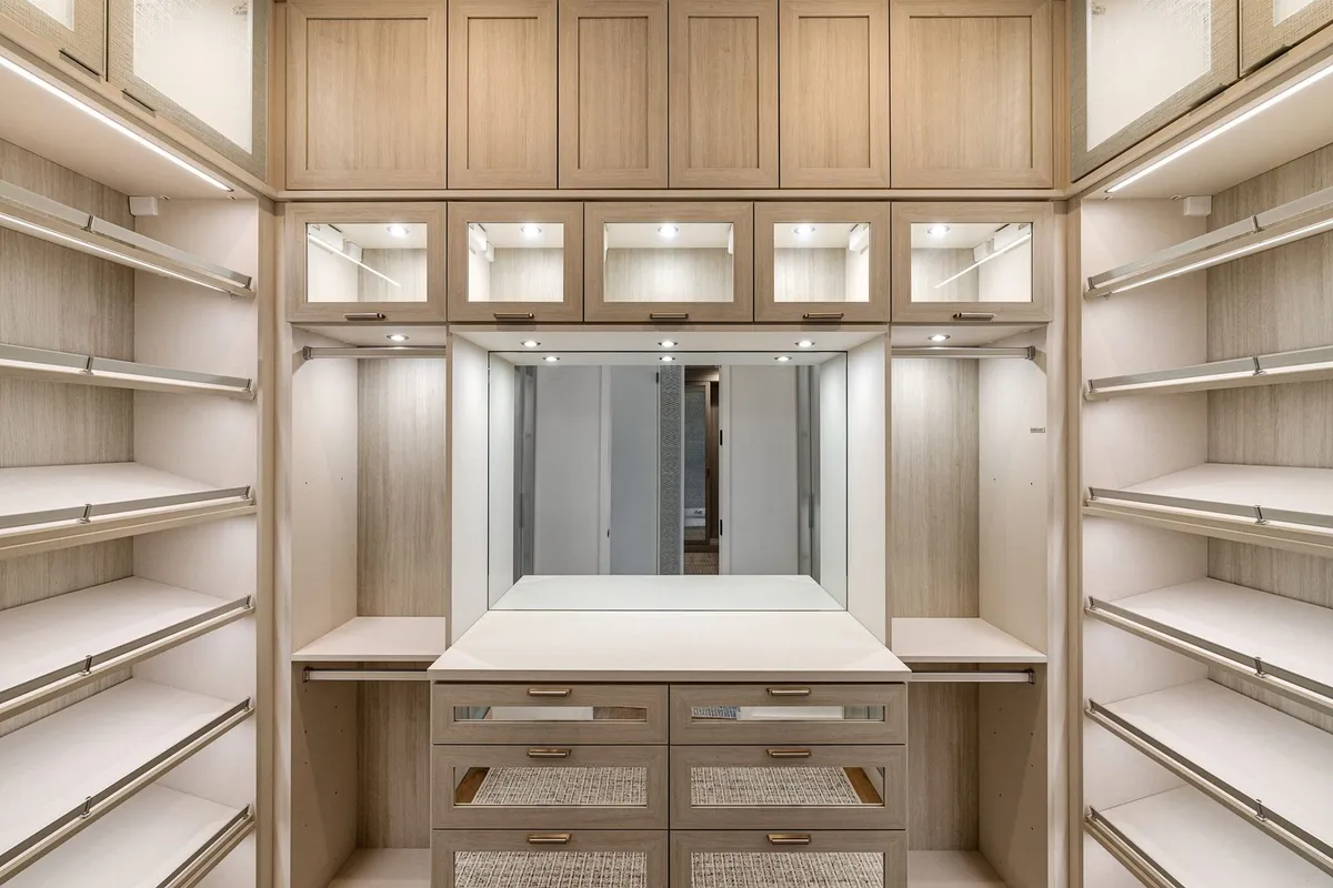 A walk-in closet with lit open shelving, glass-front upper doors and a central dresser